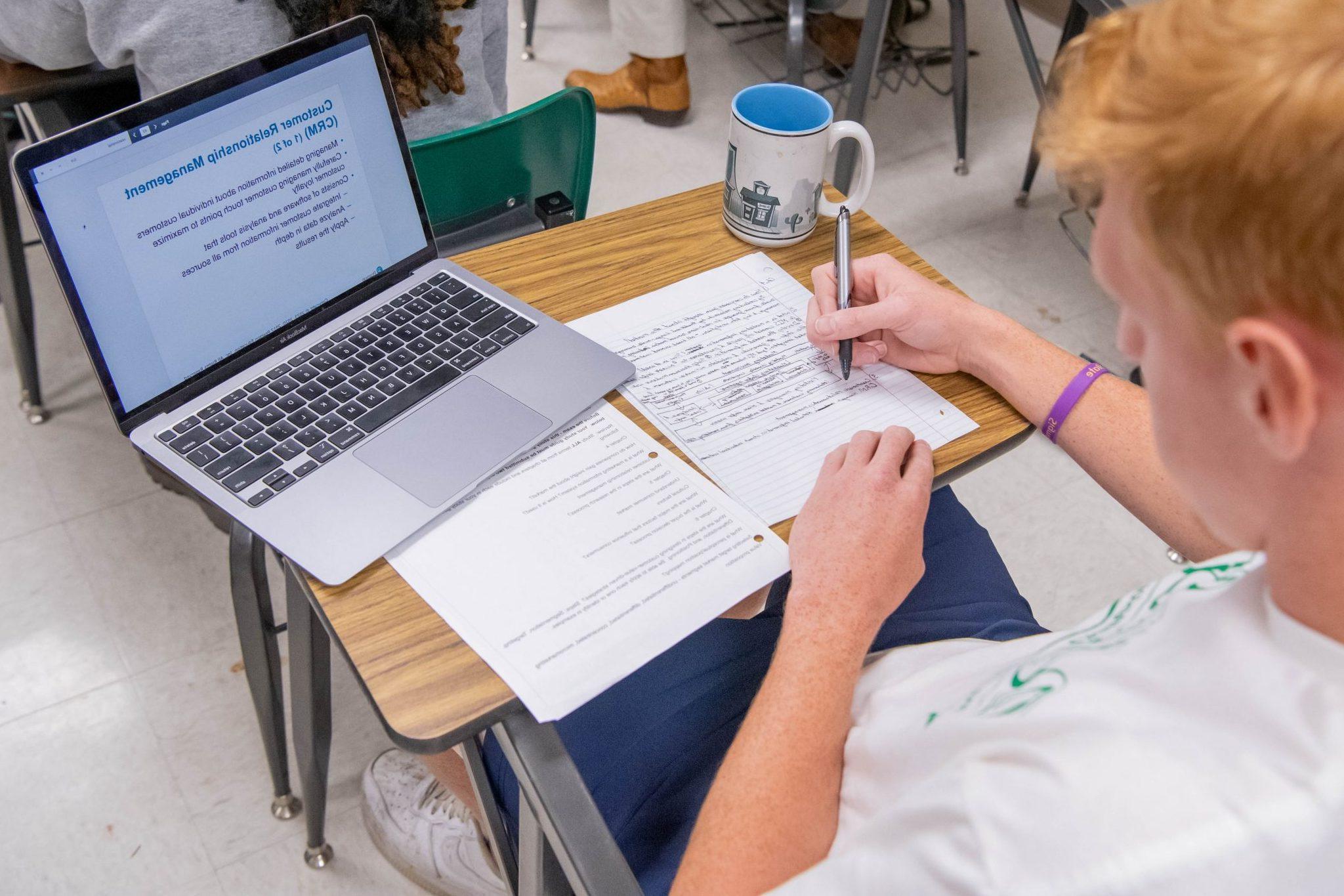 Student sitting at a desk in a classroom, taking notes on paper, 看着笔记本电脑上打开的ppt.